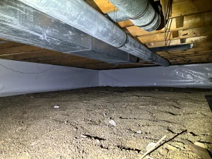 Crawl space with exposed soil and overhead metal ducts. Wooden beams support the ceiling, lined with white insulation, creating an industrial tone.