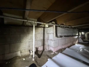 Dimly lit crawl space with exposed pipes, concrete walls, and wooden beams. Some parts covered with white plastic sheeting. Utilitarian and damp.