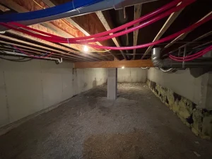 Dimly lit crawlspace with exposed wooden beams and colorful piping on the ceiling. Gravel floor and concrete walls create a raw, unfinished look.