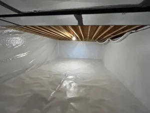 A clean, bright crawl space with white plastic lining on the floor and walls. The ceiling shows exposed wooden beams and a single light bulb.
