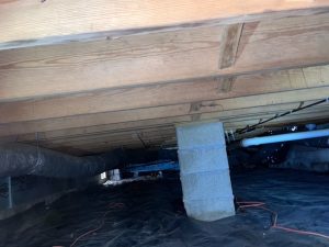 Crawl space under a house with wooden beams and a support pillar, featuring exposed pipes and ducts. The ground is covered with black plastic sheeting.
