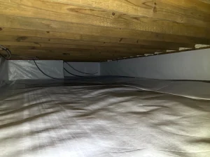 A wooden ceiling crawl space with white vapor barrier lining the floor and walls. Dim lighting and exposed cables create a utilitarian feel.